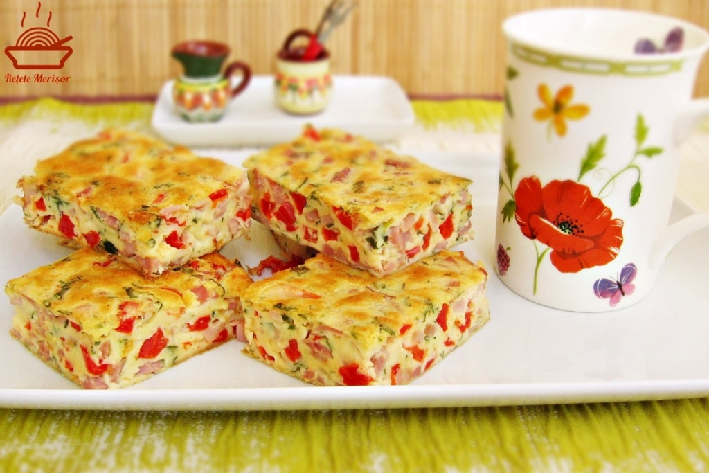Budincă aperitiv cu șuncă și ardei porționată pe un platou alb, alături de o cană și recipiente rustice